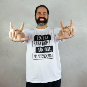 Marcelo, homem adulto de pele branca, com cabelo castanho-escuro curto e barba grisalha, está com as mãos mostrando os sinais "I love you" e sorri para a câmera. Ele veste uma camiseta branca com estampa preta, onde se lê: “Legenda para quem não ouve, mas se emociona”. O fundo da imagem é uma parede clara com acabamento em cimento queimado.