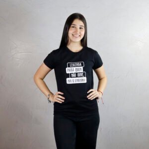 Maria Fernanda é moça jovem, magra, alta, de pele branca, cabelo longo, preto e liso, sorri para a câmera. Ela veste uma camiseta baby look preta com estampa branca escrita “Legenda para quem não ouve, mas se emociona!” e usa calça preta. O fundo da imagem é uma parede clara com acabamento de cimento queimado e ambiente bastante iluminada.
