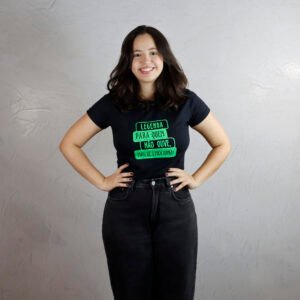 Rafaela, uma jovem de pele branca, com cabelo castanho-escuro até os ombros, sorri para a câmera. Ela veste uma camiseta baby look preta com estampa verde flúor escrita "Legenda para quem não ouve, mas se emociona" e usa calça jeans preta. Ao fundo, há uma parede clara com acabamento em cimento queimado
