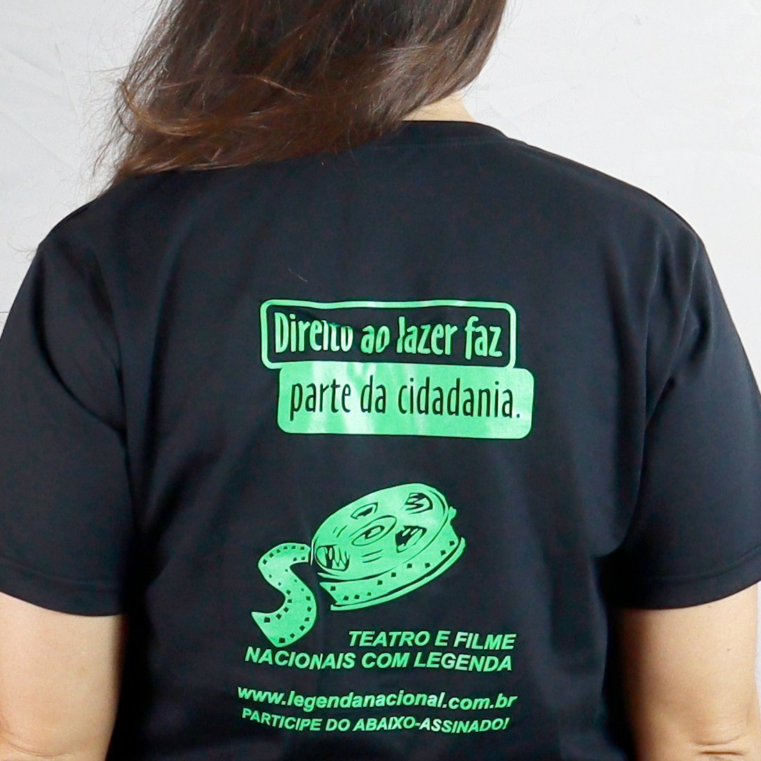 Carolina é vista de costas, em plano aproximado, em um ambiente bem iluminado, usando uma camiseta básica preta. Nas costas da camiseta há uma estampa em verde flúor. Na parte superior, em dois blocos retangulares, lê-se: ‘Direito ao lazer faz parte da cidadania.’ Abaixo do texto, há a ilustração de um rolo de filme de cinema desenrolando. Mais abaixo, aparece a frase: ‘Teatro e filme nacionais com legenda’. Na parte inferior da estampa estão o site ‘www.legendanacional.com.br’ e a frase ‘Participe do abaixo-assinado!’.