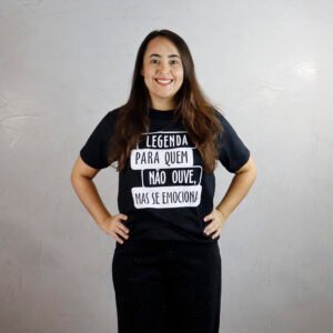 Carolina está em pé, com cabelos longos e escuros, sorrindo para a frente. Ela veste uma camiseta básica preta com estampa branca composta por quatro faixas retangulares e desalinhadas, com a frase ‘LEGENDA PARA QUEM NÃO OUVE, MAS SE EMOCIONA!’. Usa calça preta e mantém as mãos na cintura. O fundo é uma parede clara com acabamento em cimento queimado, em ambiente bem iluminado.