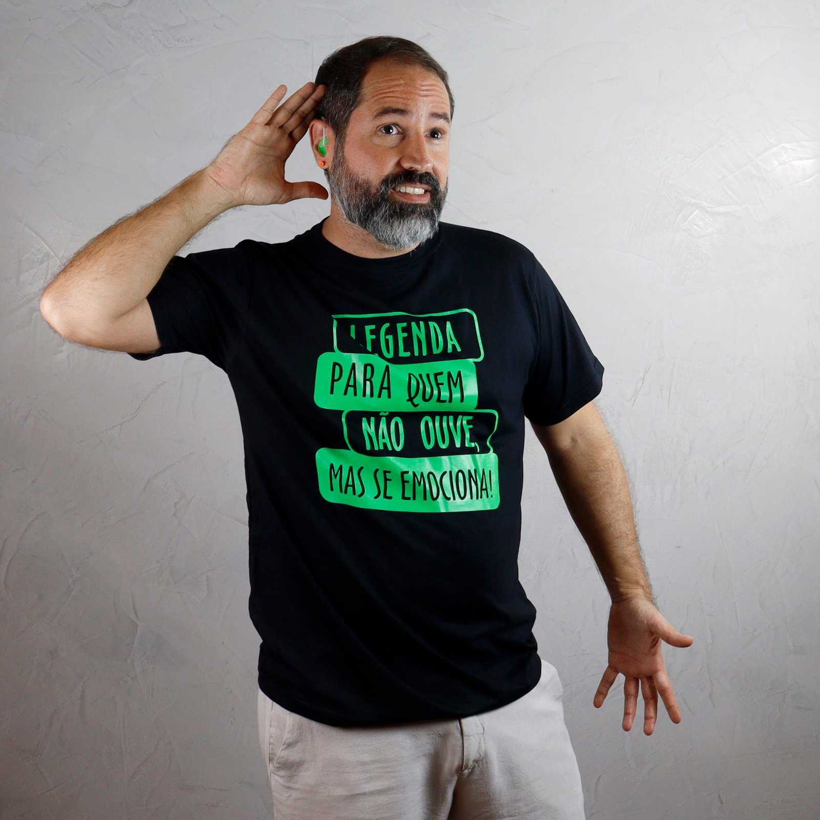 Marcelo, adulto de pele branca, barba grisalha e cabelos curtos escuros, veste camiseta preta e bermuda clara. Ele leva a mão direita atrás da orelha, como se estivesse tentando ouvir, enquanto a mão esquerda permanece aberta, voltada para baixo. A camiseta traz estampa verde fluorescente com quatro faixas retangulares horizontais e desalinhadas, formando a frase ‘Legenda para quem não ouve, mas se emociona!’. Marcelo usa aparelho auditivo verde-flúor na orelha direita. O fundo é uma parede clara de cimento queimado.