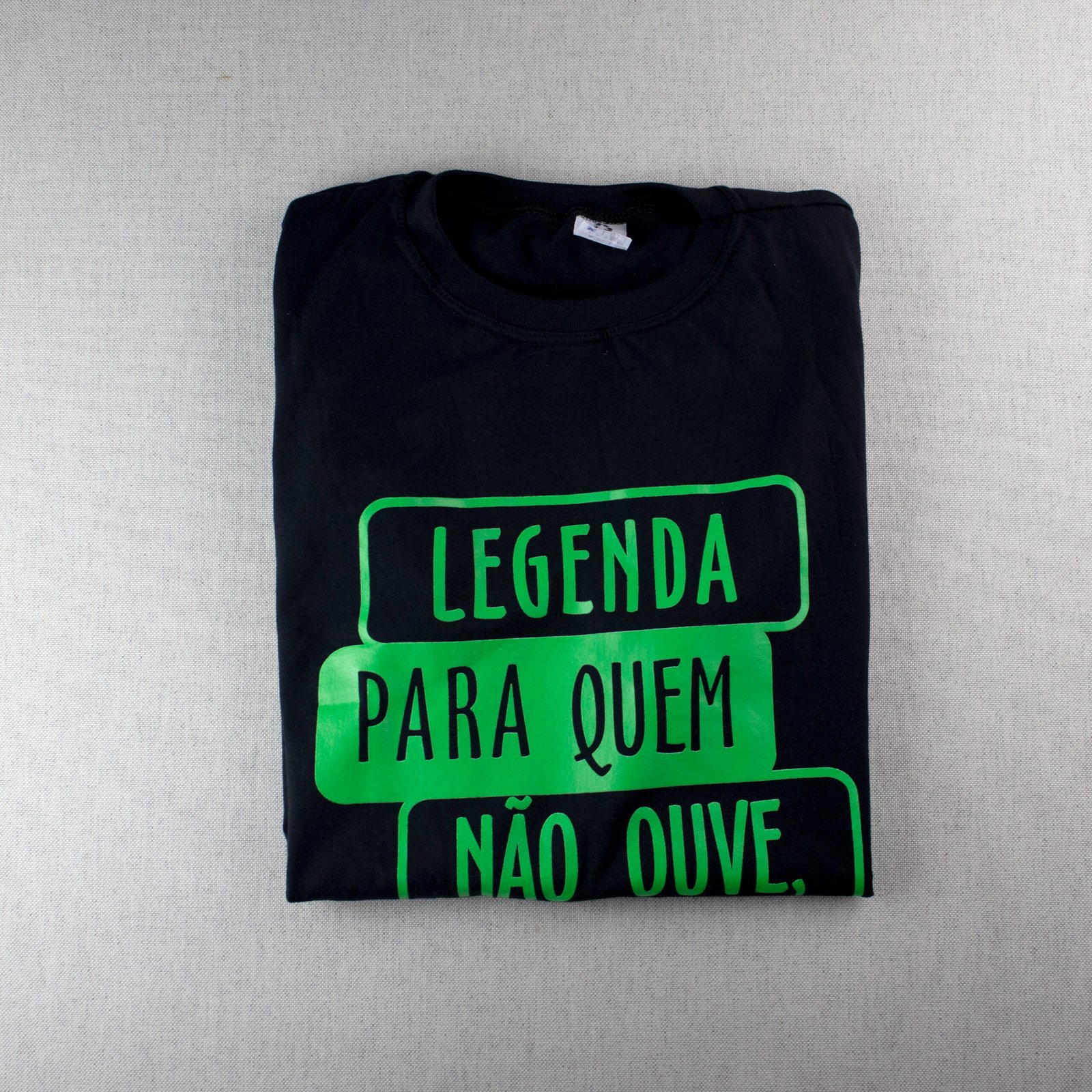 Na imagem quadrada há uma camisa básica preta dobrada em fundo branco. A estampa verde flúor forma blocos retangulares com a frase incompleta: "Legenda para quem não ouve,". O primeiro bloco contendo "legenda" possui letras verdes em fundo preto. O segundo bloco, contendo "para quem", está logo abaixo, com letras pretas em fundo verde flúor. E o terceiro bloco: "não ouve," possui letras verdes em fundo preto.