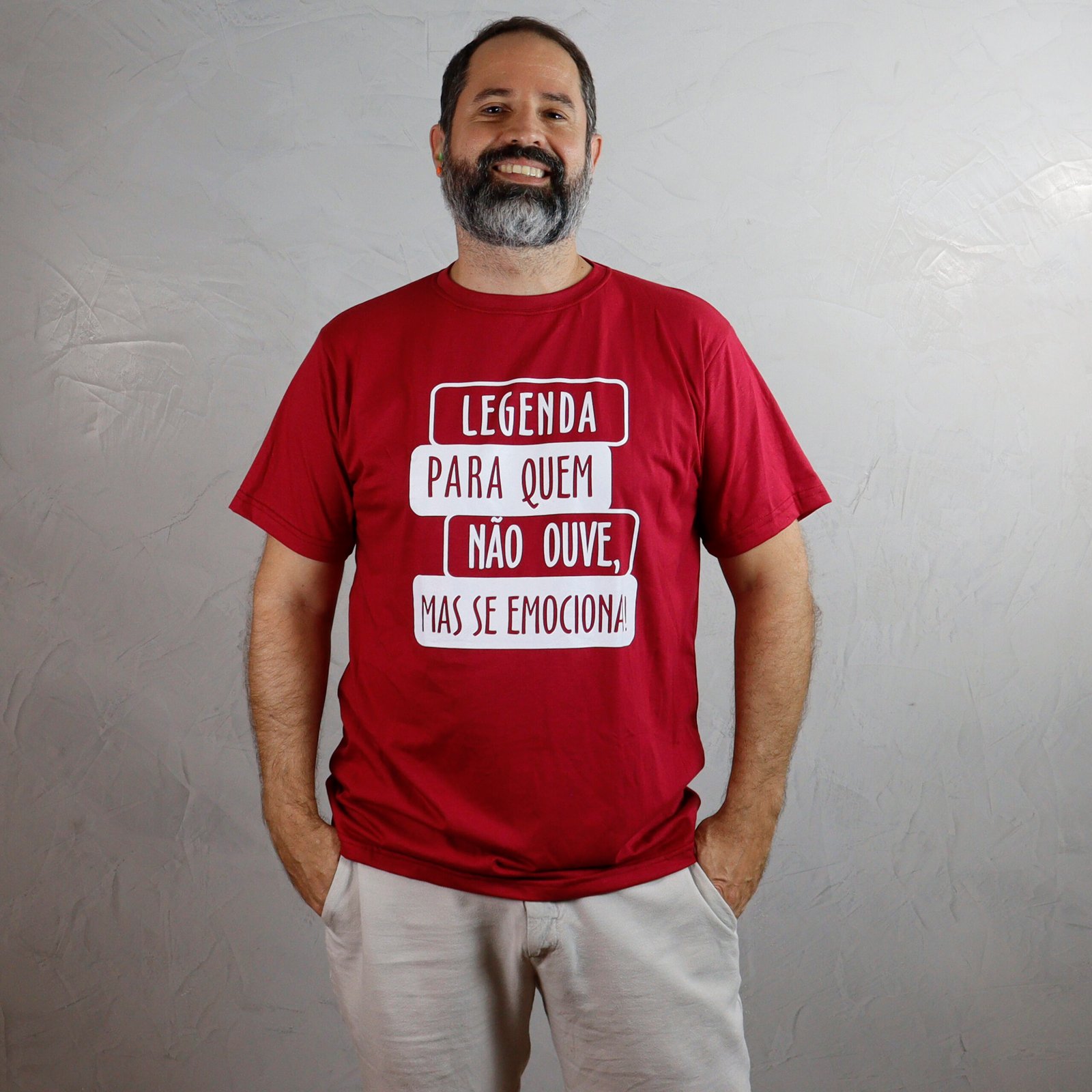 Marcelo, homem adulto de pele branca, com cabelo castanho-escuro curto e barba grisalha, está com as mãos nos bolsos e sorri para a câmera. Ele veste uma camiseta vermelha com estampa branca, onde se lê: “Legenda para quem não ouve, mas se emociona”. O fundo da imagem é uma parede clara com acabamento em cimento queimado.