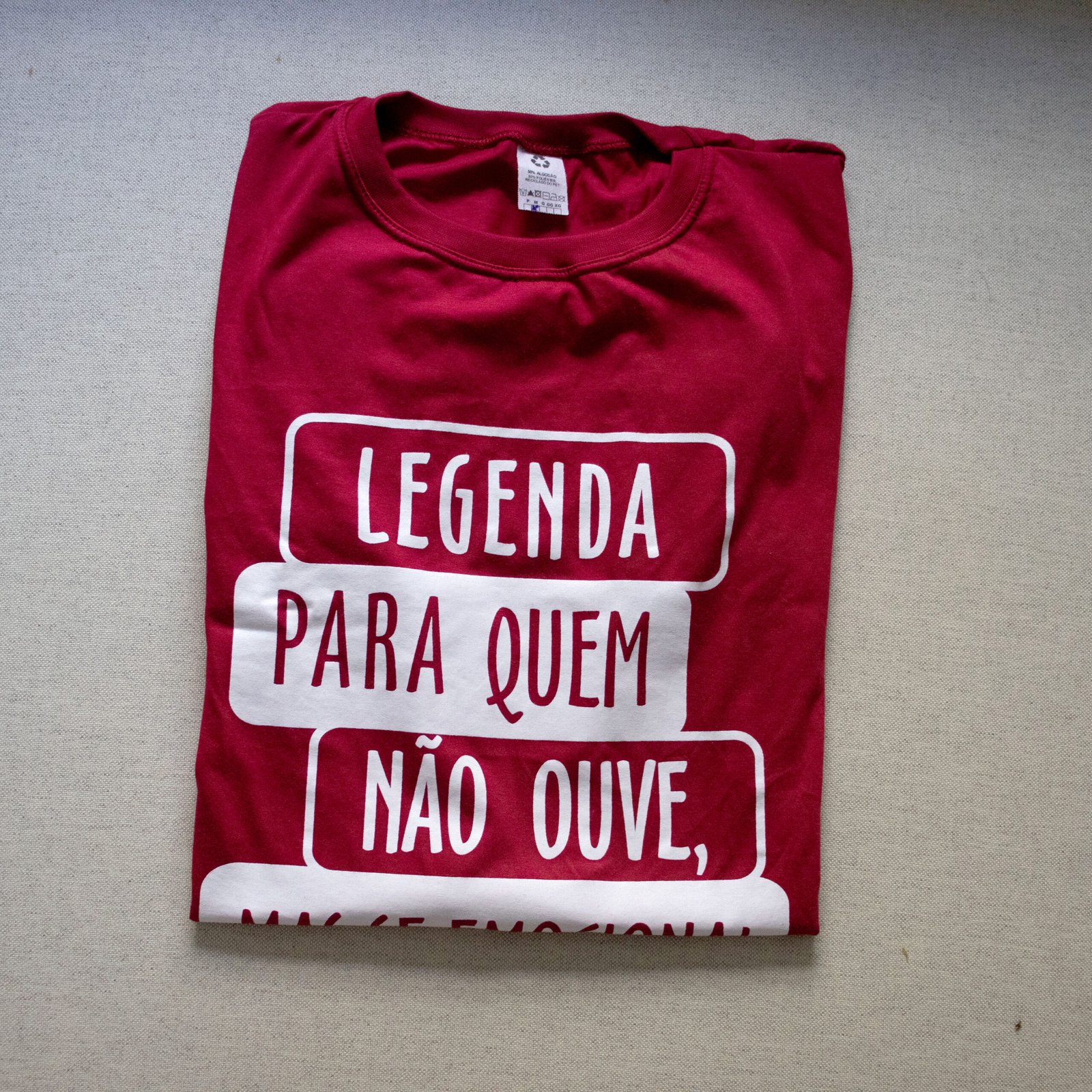 Na imagem quadrada há uma camisa vermelha dobrada em fundo branco. A estampa branca forma blocos retangulares com a frase incompleta: "Legenda para quem não ouve". O primeiro e o terceiro blocos, contendo "legenda" e "não ouve", possuem letras brancas em fundo vermelho. O segundo bloco, contendo "para quem", está logo abaixo, com letras vermelhas em fundo branco.