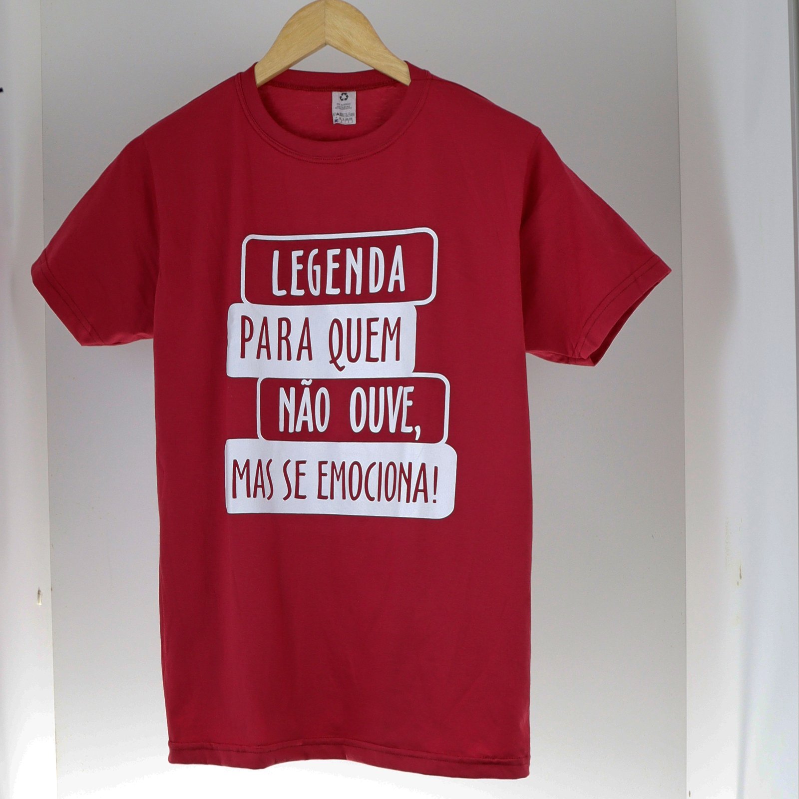 Uma camisa básica de cor vermelha escura, de mangas curtas e gola redonda, é vista de frente, pendurada em um cabide de madeira clara. A peça está sobre um fundo branco liso. No centro da camisa, há uma estampa em letras brancas, dispostas em três linhas, cada uma envolta em um contorno retangular branco arredondado, como se fossem legendas de tela: Primeira linha: "LEGENDA" Segunda linha: "PARA QUEM" Terceira linha: "NÃO OUVE," Quarta linha: "MAS SE EMOCIONA!"