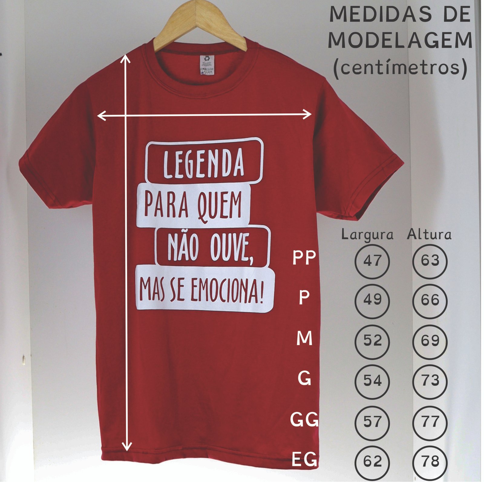 Camiseta vermelha pendurada em um cabide de madeira, fotografada de frente sobre fundo claro. Na estampa branca, lê-se: “Legenda para quem não ouve, mas se emociona!”. Setas indicam as medidas da peça: largura horizontal e altura vertical. À direita, há uma tabela de medidas de modelagem em centímetros para os tamanhos PP, P, M, G, GG e XG, mostrando larguras entre 47 e 62 cm e alturas entre 63 e 78 cm.