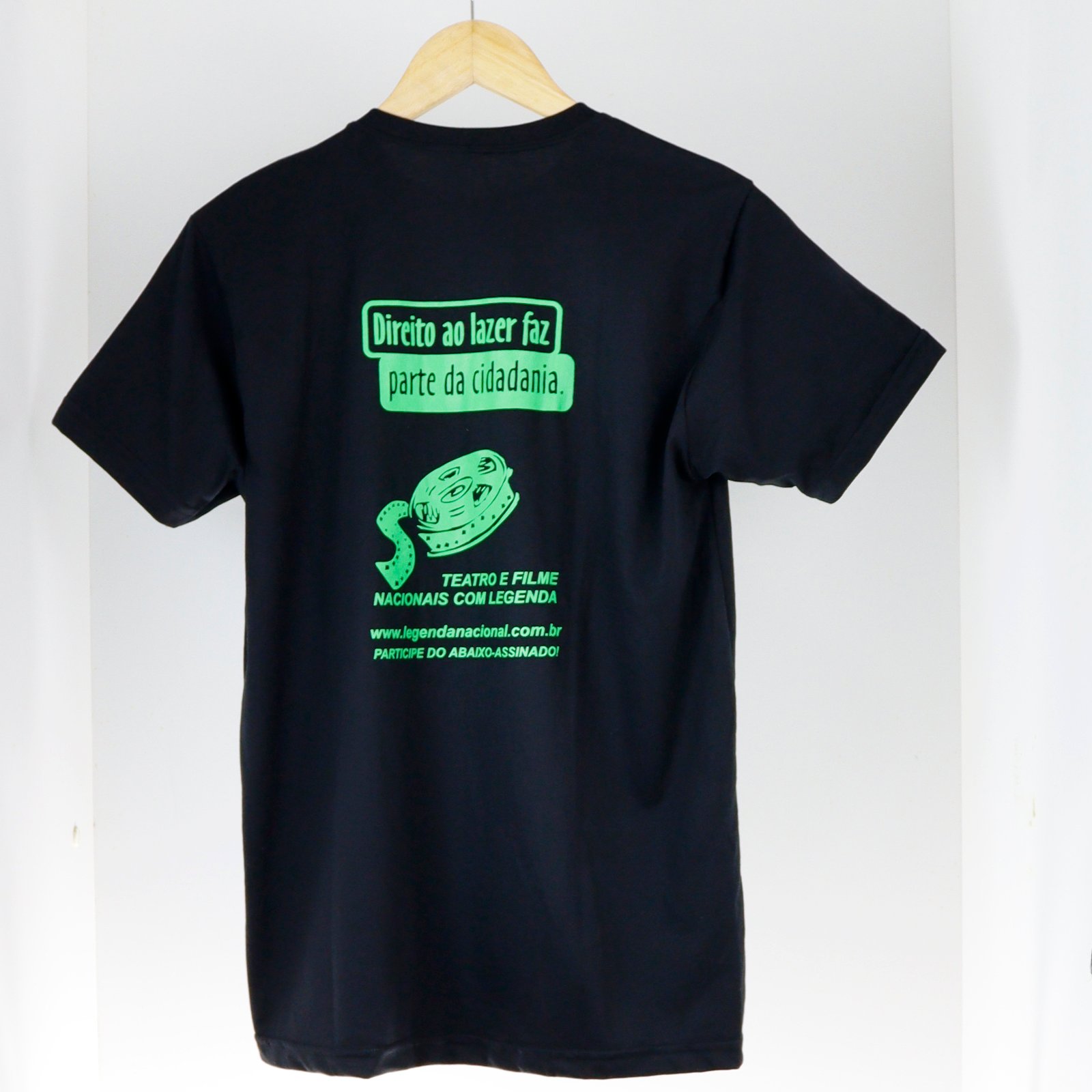Camiseta básica preta pendurada em um cabide de madeira, fotografada de frente sobre fundo claro. Nas costas, a estampa verde flúor traz os textos ‘Direito ao lazer faz parte da cidadania’ e ‘Teatro e filme nacionais com legenda’, além da ilustração de um rolo de filme de cinema, do site ‘www.legendanacional.com.br’ e da frase ‘Participe do abaixo-assinado!’.