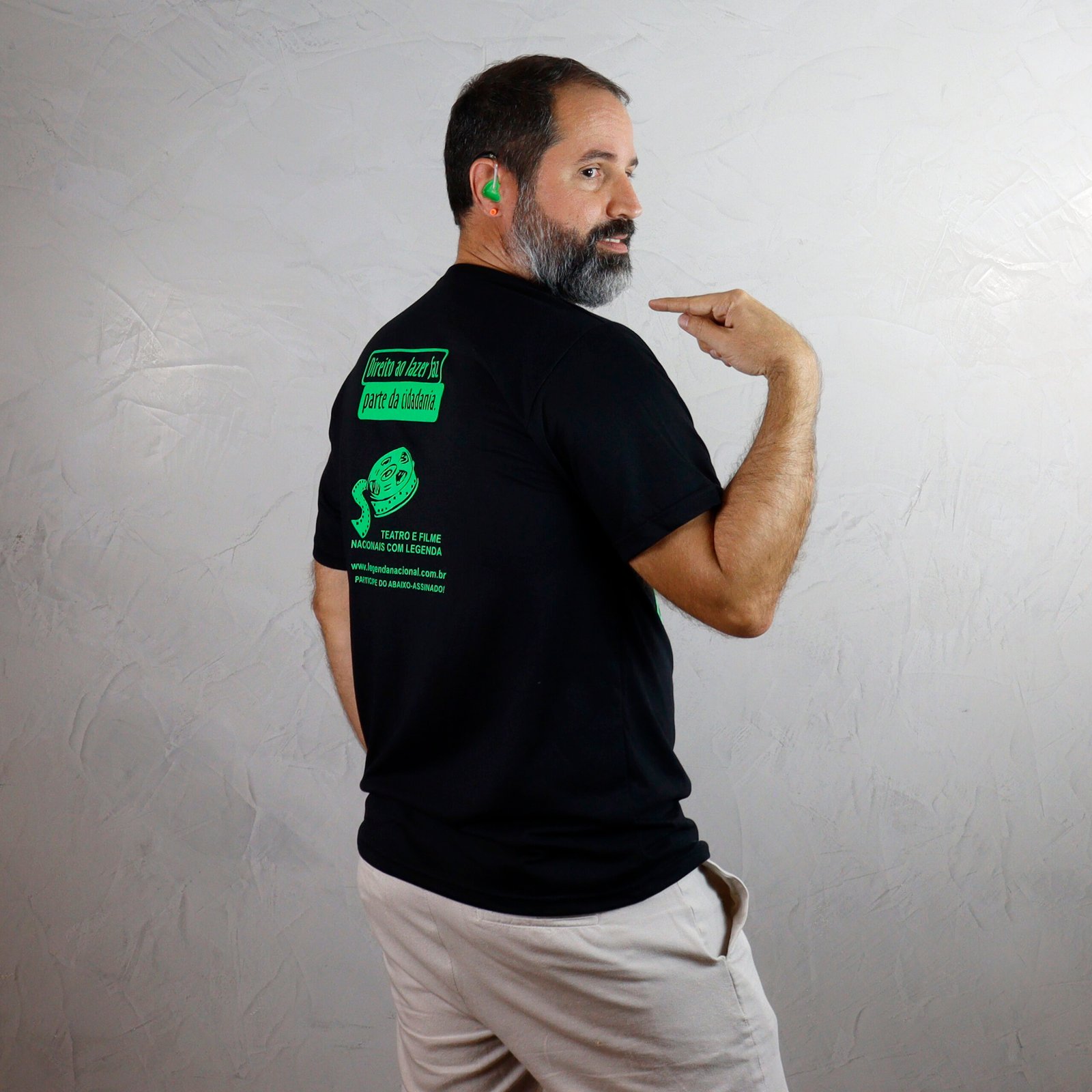 Marcelo, adulto de pele branca, barba grisalha e cabelos curtos escuros, está de costas, voltado para a direita. Ele veste camiseta básica preta e bermuda clara e usa um aparelho auditivo verde na orelha. Com a mão direita, aponta para a estampa verde flúor nas costas da camiseta, com os textos ‘Direito ao lazer faz parte da cidadania’ e ‘Teatro e filme nacionais com legenda’, além do desenho de uma película de cinema. O fundo é uma parede cinza-clara com acabamento em cimento queimado.