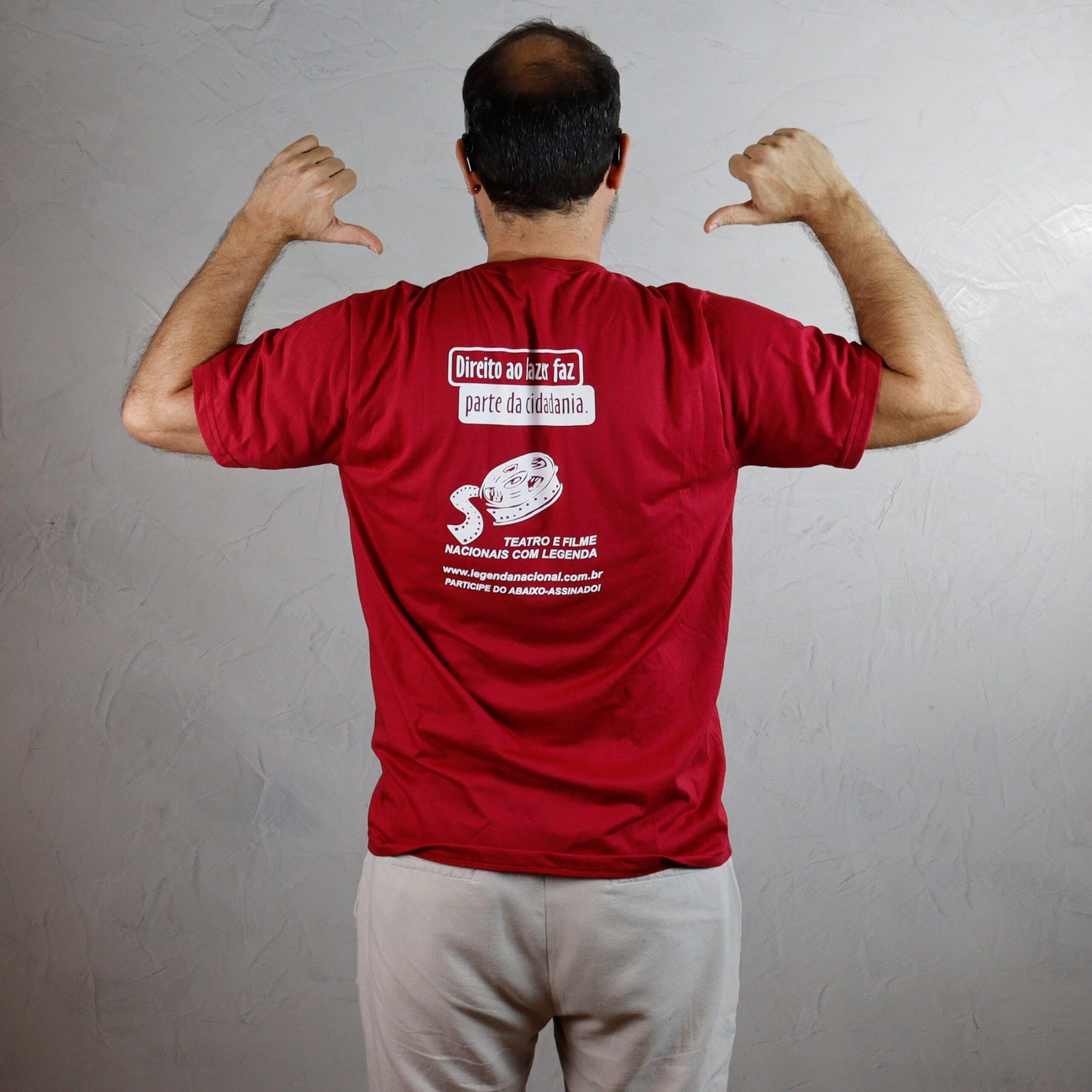 Um homem branco de costas, com calças cinzas claras e uma camisa básica vermelha escura. Ele tem cabelo escuro e está ligeiramente calvo no topo. O homem faz gestos de "jóia" ou "legal" com os polegares para baixo, apontando para si mesmo. Na parte de trás da camisa, há uma estampa branca com texto e um desenho: Topo (em uma caixa branca): "Direito ao lazer faz parte da cidadania." Abaixo: Desenho de uma fita de filme em espiral. Mais abaixo: "TEATRO É FILME NACIONAIS COM LEGENDA" Fundo: "WWW.LEGENDANACIONAL.COM.BR / PARTICIPE DO ABAIXO-ASSINADO!"
