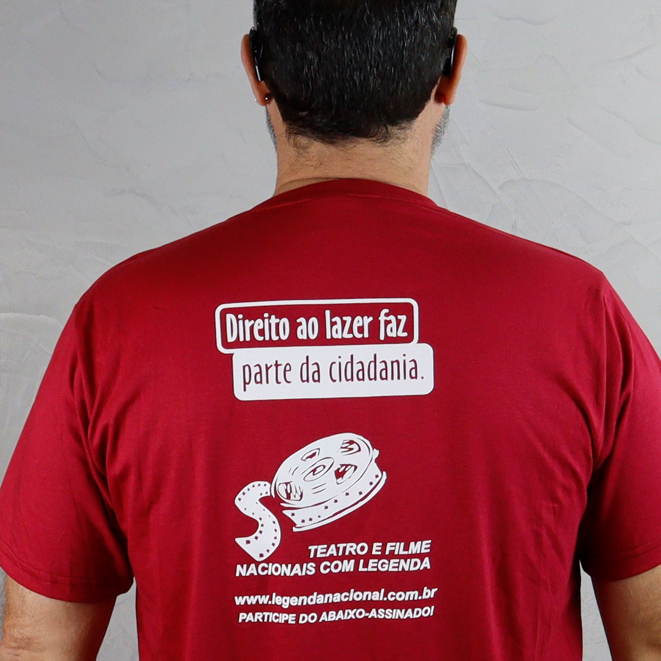 Pessoa vista de costas usando camiseta vermelha, em ambiente interno com fundo claro. Na parte superior das costas há uma estampa branca com a frase: “Direito ao lazer faz parte da cidadania.” Abaixo, aparece uma ilustração de um rolo de filme de cinema com expressão facial estilizada. Logo abaixo, lê-se: “Teatro e filme nacionais com legenda”. Na parte inferior da estampa, consta o site “www.legendanacional.com.br” e a frase “Participe do abaixo-assinado!”.