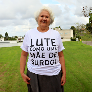 A imagem mostra uma mulher idosa sorridente, de pele clara, posicionada de frente no centro do enquadramento. Ela está em um ambiente externo, com um gramado verde e céu nublado ao fundo. Vestimenta: Ela veste uma camiseta branca de mangas curtas. Estampa da Camiseta: O texto está centralizado no peito em letras grandes, pretas e em caixa alta: LUTE COMO UMA MÃE DE SURDO! Aparência Física: A mulher tem cabelos brancos curtos e ondulados. Seu sorriso é largo e demonstra entusiasmo. Cenário: Ao fundo, vê-se um campo gramado, algumas árvores à direita e construções urbanas ao longe sob um céu com muitas nuvens claras.