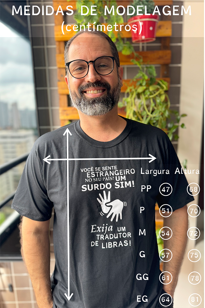 Descrição da Imagem: Fotografia vertical de Marcelo Pedrosa, adulto sorridente, com barba e bigode grisalhos e óculos de armação preta, posando numa varanda de apartamento. Ele veste uma t-shirt preta com estampa branca no peito e tem as mãos nos bolsos das calças. No topo da imagem, lê-se em letras maiúsculas brancas: "MEDIDAS DE MODELAGEM (centímetros)". Sobre o peito do homem, há duas setas brancas cruzadas que indicam a forma de medição: Uma seta vertical longa que vai da gola até à bainha inferior (indicando a Altura). Uma seta horizontal curta na região do tórax (indicando a Largura). A estampa da t-shirt contém a frase: "VOCÊ SE SENTE ESTRANGEIRO NO SEU PAÍS? UM SURDO SIM!", acompanhada pela ilustração de duas mãos abertas em sinal de Libras e, abaixo, o texto "Exija UM TRADUTOR DE LIBRAS!"