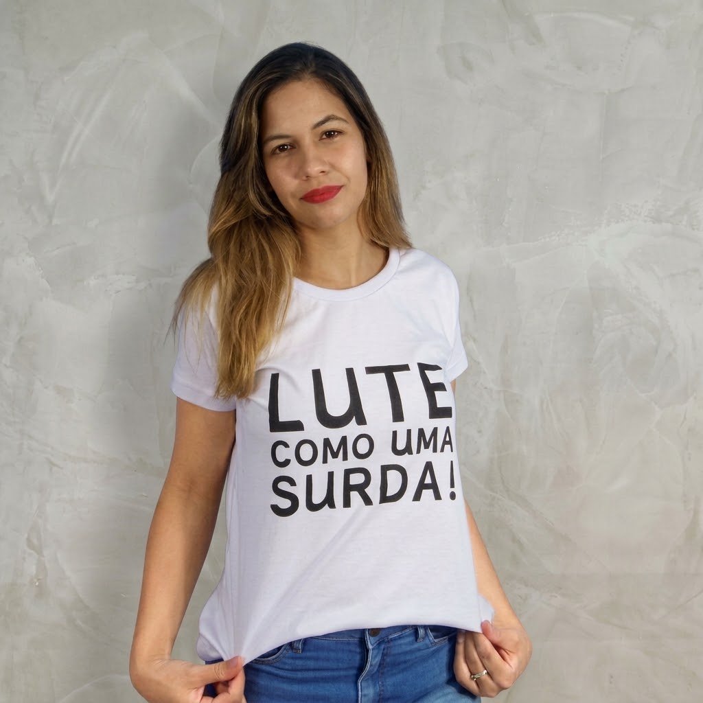 Letícia Lima com a camiseta branca: Mulher de pele branca, cabelos liso, batom vermelho vestindo uma camiseta baby look branca com a estampa preta LUTE COMO UMA SURDA! e short jeans azul, posando em frente a uma parede cinza.