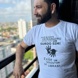 Alan, adulto de barba escura e curta está sorrindo. Ele está com camiseta branca que está vestindo. A camiseta tem a frase "LUTE COMO UM SURDO!" estampada em letras pretas e maiúsculas. O homem está voltado para uma sacada com a vista de prédios da cidade. Sua expressão é alegre e orgulhosa.