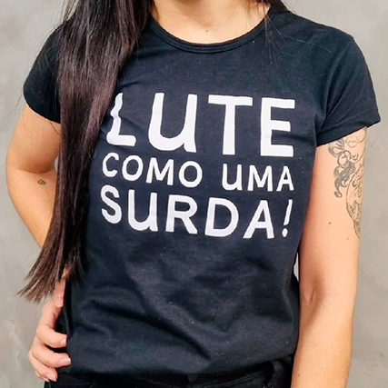 Descrição da imagem #ParaTodosVerem: Ao fundo, uma parede cinza de cimento queimado. Fabiane está no centro, em pé. Ela tem pele branca e cabelo liso, castanho escuro, que vai até a cintura. Fabiane sorri e apoia a mão direita na cintura. Ela veste uma blusa preta com a estampa branca que diz: "Lute como uma surda.