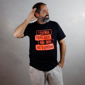 Marcelo, adulto de pele branca, barba grisalha e cabelos curtos escuros, veste camiseta preta e bermuda clara. Ele leva a mão direita atrás da orelha, como se estivesse tentando ouvir, enquanto a mão esquerda no bolso. A camiseta traz estampa laranja néon com quatro faixas retangulares horizontais e desalinhadas, formando a frase ‘Legenda para quem não ouve, mas se emociona!’. Marcelo usa aparelho auditivo com molde de silicone da cor laranja néon na orelha direita. O fundo é uma parede clara de cimento queimado.