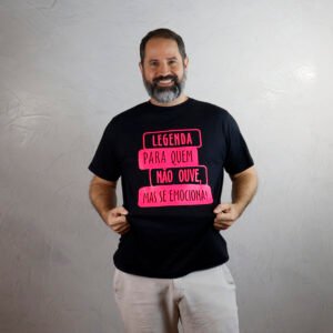 Marcelo, homem adulto de pele branca, cabelo castanho escuro curto e barba grisalha, sorri para a câmera. Ele veste uma camiseta básica preta com estampa rosa néon escrita “Legenda para quem não ouve, mas se emociona”. O fundo da imagem é uma parede clara com acabamento de cimento queimado.